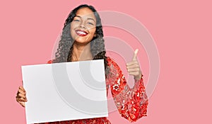 Young african american girl holding blank empty banner smiling happy and positive, thumb up doing excellent and approval sign