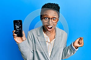 Young african american girl holding broken smartphone showing cracked screen screaming proud, celebrating victory and success very