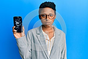 Young african american girl holding broken smartphone showing cracked screen thinking attitude and sober expression looking self