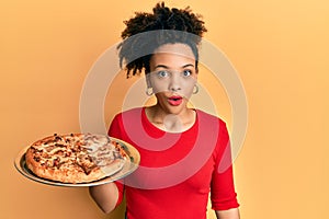 Young african american girl holding italian pizza scared and amazed with open mouth for surprise, disbelief face