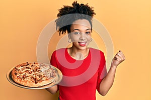 Young african american girl holding italian pizza screaming proud, celebrating victory and success very excited with raised arm