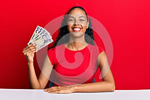 Young african american girl holding japanese yen banknotes sitting on the table looking positive and happy standing and smiling