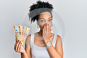 Young african american girl holding philippine peso banknotes covering mouth with hand, shocked and afraid for mistake