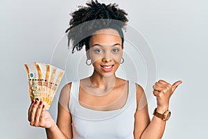 Young african american girl holding philippine peso banknotes pointing thumb up to the side smiling happy with open mouth