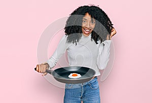 Young african american girl holding skillet with fried egg screaming proud, celebrating victory and success very excited with