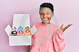 Young african american girl holding sweet pastries in a box celebrating achievement with happy smile and winner expression with photo