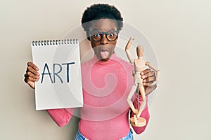 Young african american girl holding wooden manikin and art word on notebook sticking tongue out happy with funny expression
