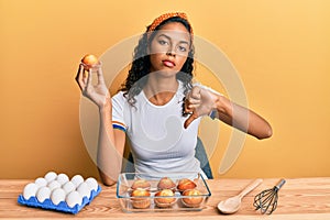 Young african american girl making muffins sitting on the table with angry face, negative sign showing dislike with thumbs down,