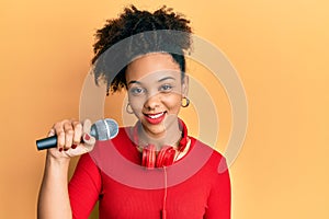 Young african american girl singing song using microphone and headphones looking positive and happy standing and smiling with a
