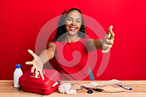 Young african american girl sitting on the table with first aid kit making cure looking at the camera smiling with open arms for