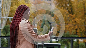 Young african american girl standing with phone and coffee on autumn day outdoors. Student waiting for meeting, female