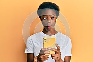 Young african american girl using smartphone and earphones making fish face with mouth and squinting eyes, crazy and comical