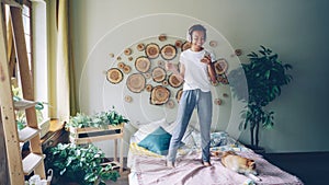 Young African American girl is using smartphone, listening to music with headphones and jumping on bed enjoying free