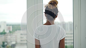 Young African American girl is walking to the window at home and parting the curtains then enjoying view from the window