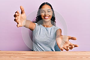 Young african american girl wearing casual clothes sitting on the table looking at the camera smiling with open arms for hug