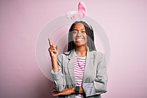 Young african american girl wearing cute easter bunny ears over pink background with a big smile on face, pointing with hand and