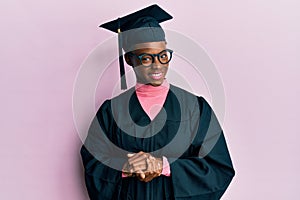 Young african american girl wearing graduation cap and ceremony robe with hands together and crossed fingers smiling relaxed and