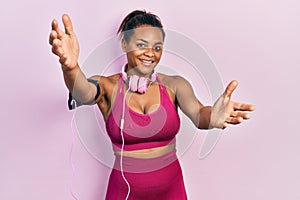 Young african american girl wearing gym clothes and using headphones looking at the camera smiling with open arms for hug