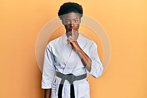 Young african american girl wearing karate kimono and black belt asking to be quiet with finger on lips