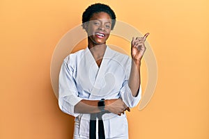 Young african american girl wearing karate kimono and black belt with a big smile on face, pointing with hand and finger to the