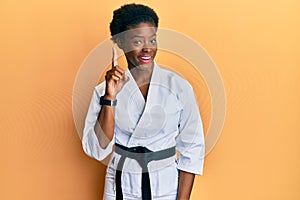 Young african american girl wearing karate kimono and black belt showing and pointing up with finger number one while smiling