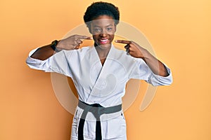 Young african american girl wearing karate kimono and black belt smiling cheerful showing and pointing with fingers teeth and