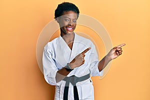 Young african american girl wearing karate kimono and black belt smiling and looking at the camera pointing with two hands and