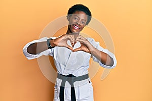 Young african american girl wearing karate kimono and black belt smiling in love doing heart symbol shape with hands