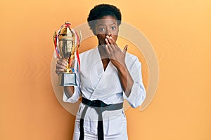 Young african american girl wearing karate kimono holding trophy covering mouth with hand, shocked and afraid for mistake
