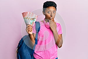 Young african american girl wearing student backpack and holding new zealand dollars covering mouth with hand, shocked and afraid