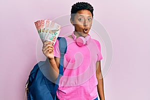 Young african american girl wearing student backpack and holding new zealand dollars scared and amazed with open mouth for