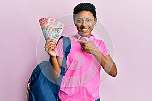 Young african american girl wearing student backpack and holding new zealand dollars smiling happy pointing with hand and finger
