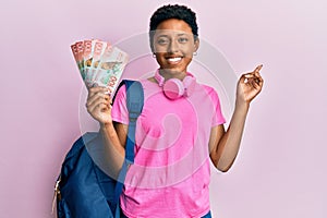 Young african american girl wearing student backpack and holding new zealand dollars smiling happy pointing with hand and finger