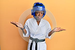 Young african american girl wearing taekwondo kimono and protection helmet clueless and confused with open arms, no idea and