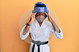 Young african american girl wearing taekwondo kimono and protection helmet with hand on head, headache because stress