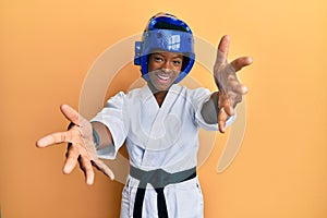 Young african american girl wearing taekwondo kimono and protection helmet looking at the camera smiling with open arms for hug