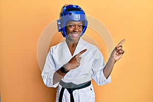 Young african american girl wearing taekwondo kimono and protection helmet smiling and looking at the camera pointing with two