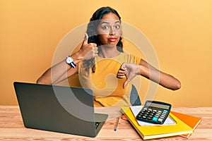 Young african american girl working at the office with laptop and calculator doing thumbs up and down, disagreement and agreement