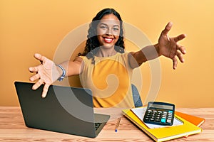 Young african american girl working at the office with laptop and calculator looking at the camera smiling with open arms for hug