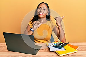 Young african american girl working at the office with laptop and calculator pointing to the back behind with hand and thumbs up,