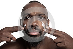 Young african-american guy applying face cream under his eyes on white background