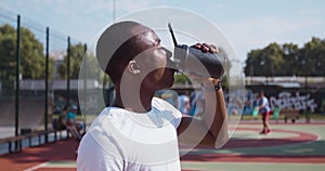 Young african american guy drinking water from sport bottle at outdoor basketball court, side view, slow motion