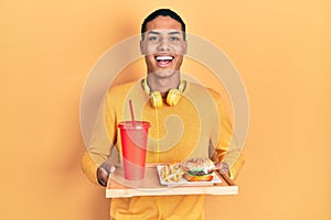 Young african american guy eating a tasty classic burger smiling and laughing hard out loud because funny crazy joke