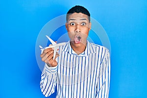 Young african american guy holding airplane toy scared and amazed with open mouth for surprise, disbelief face