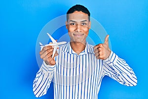 Young african american guy holding airplane toy smiling happy and positive, thumb up doing excellent and approval sign