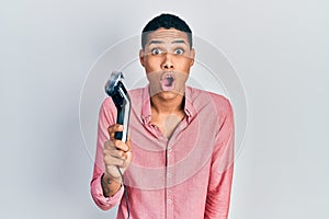 Young african american guy holding electric razor machine scared and amazed with open mouth for surprise, disbelief face