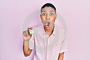 Young african american guy holding face moisturizer cream scared and amazed with open mouth for surprise, disbelief face