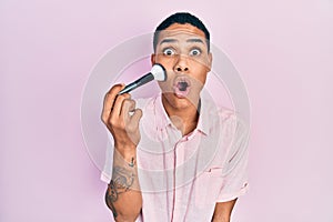 Young african american guy holding makeup brush scared and amazed with open mouth for surprise, disbelief face