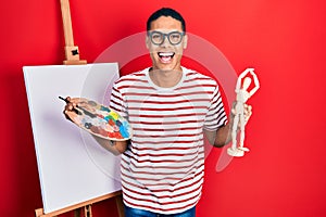 Young african american guy holding painter palette and art manikin smiling and laughing hard out loud because funny crazy joke