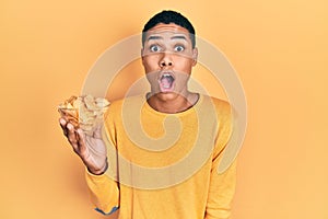 Young african american guy holding potato chip scared and amazed with open mouth for surprise, disbelief face